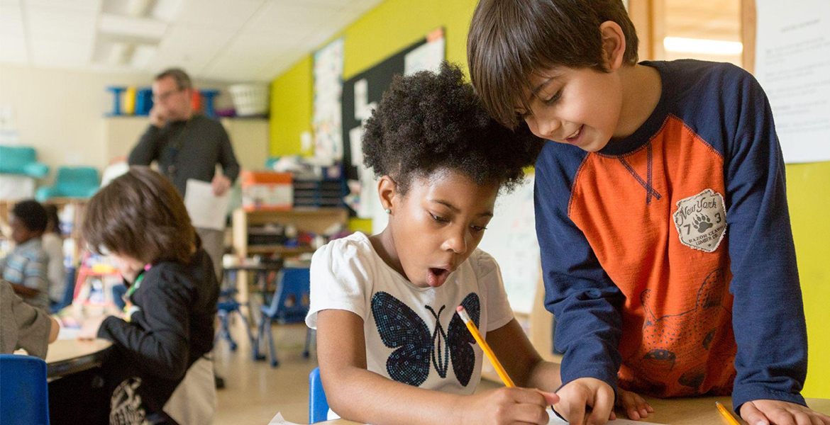 Image of two younger students writing with a pencil in a classroom